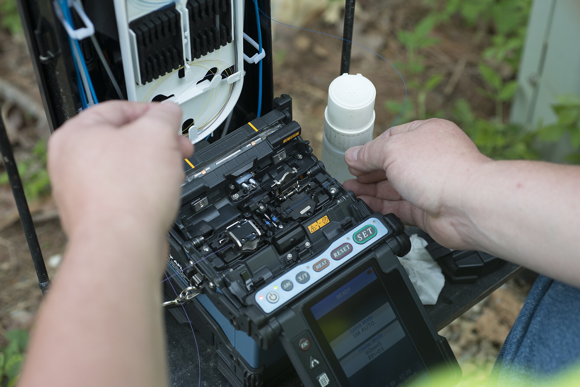 A technician's hands feeding a single optical fibre into a portable fusion splicer set up outdoors beside an open splice tray